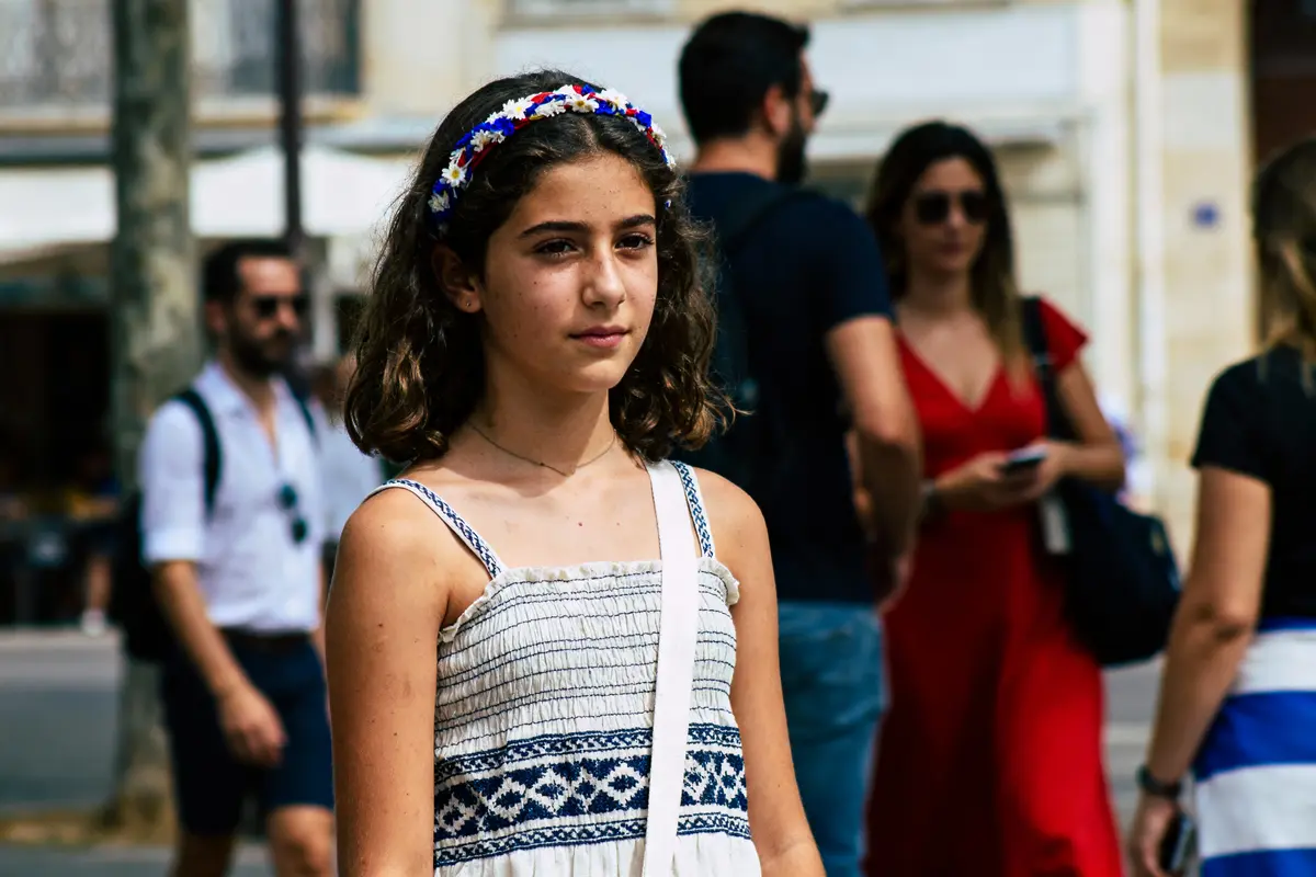 Jeune fille portant une robe estivale blanche et un bandeau fleuri, marchant en ville parmi des adultes.