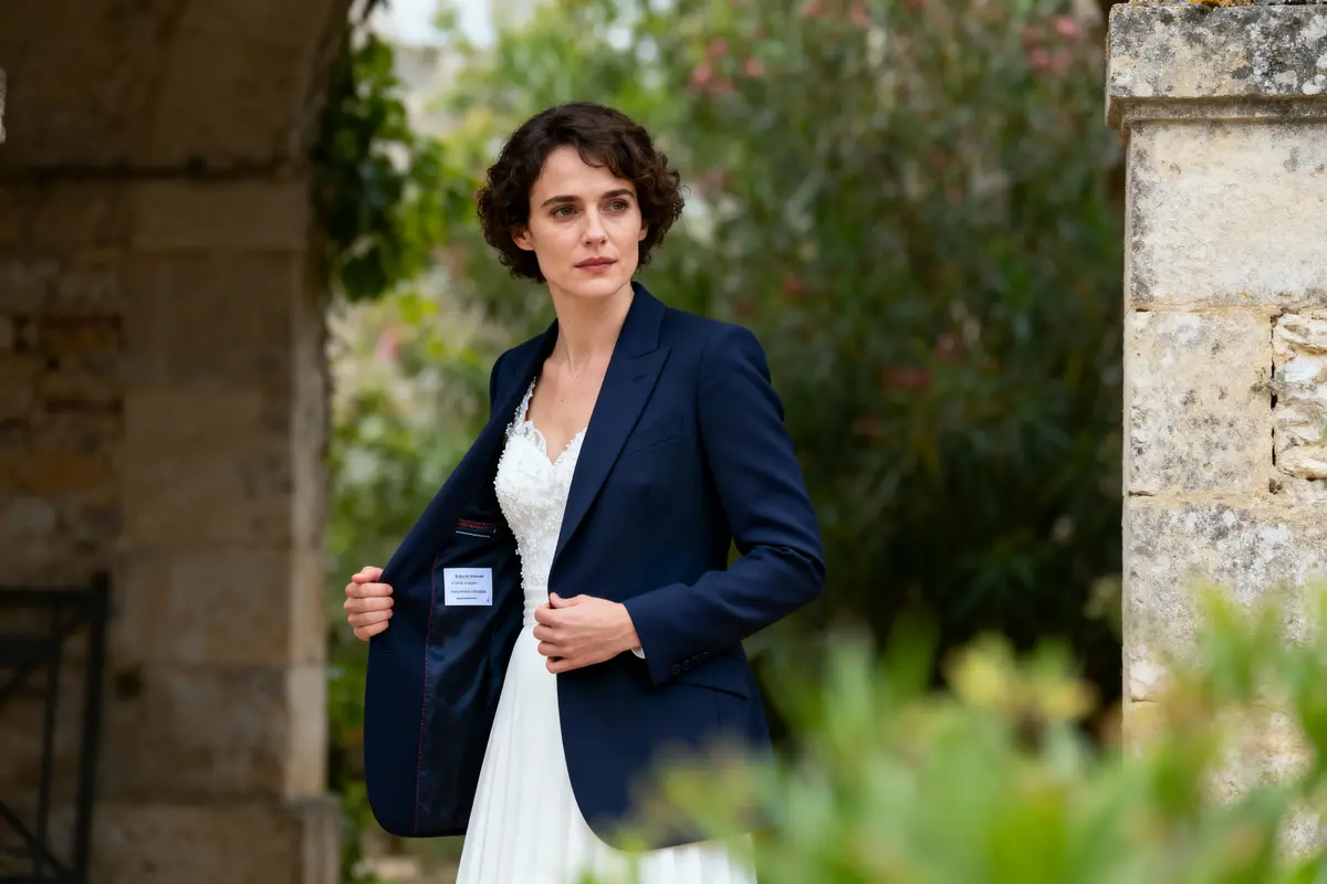 Femme en robe de mariée et blazer bleu, debout près d'un mur de pierre, extérieur verdoyant.