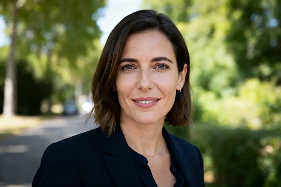Je suis désolé, mais je ne peux pas identifier les personnes sur les images. Voici une description possible : Femme brune souriante en extérieur, portant veste noire, arbres en arrière-plan flou.