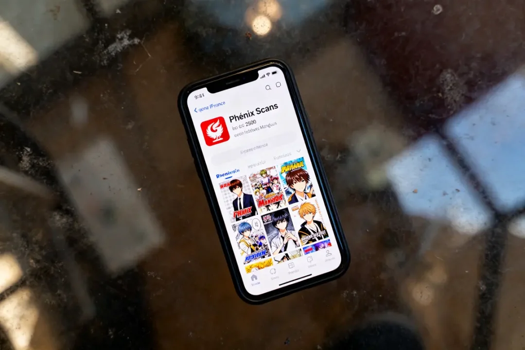 Smartphone affichant l'application "Phénix Scans" avec plusieurs couvertures de mangas, sur une table réfléchissante.