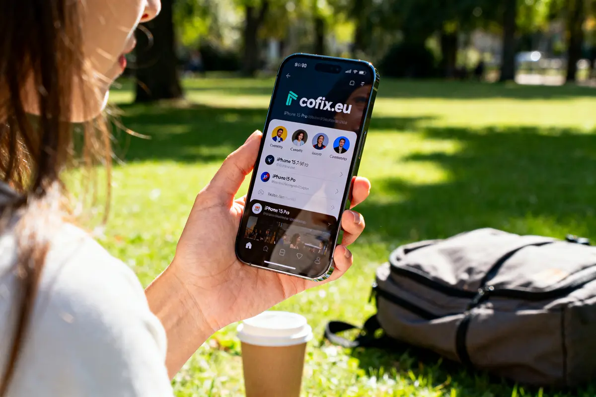 Personne tenant un smartphone dans un parc, écran affichant cofix.eu, avec sac à dos et café à proximité.