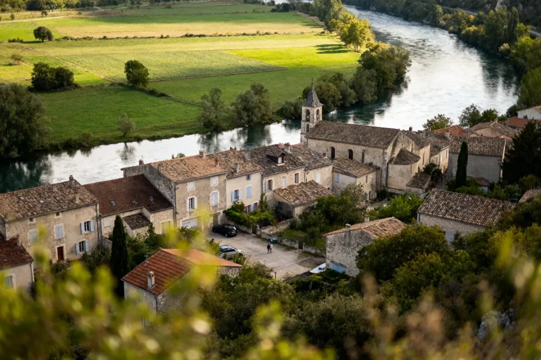 Village pittoresque près d'une rivière, maisons anciennes en pierre, champ verdoyant en arrière-plan, arbres environnants.