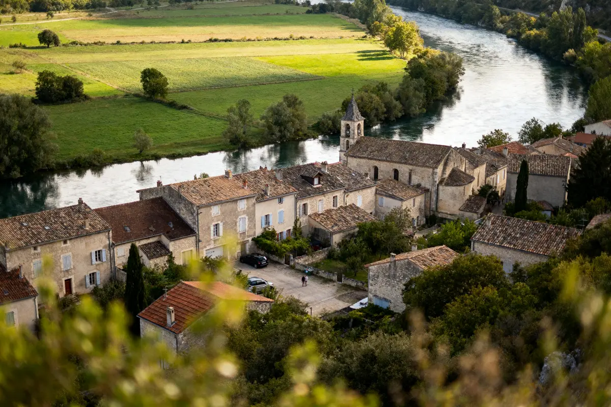 Village pittoresque près d'une rivière, maisons anciennes en pierre, champ verdoyant en arrière-plan, arbres environnants.