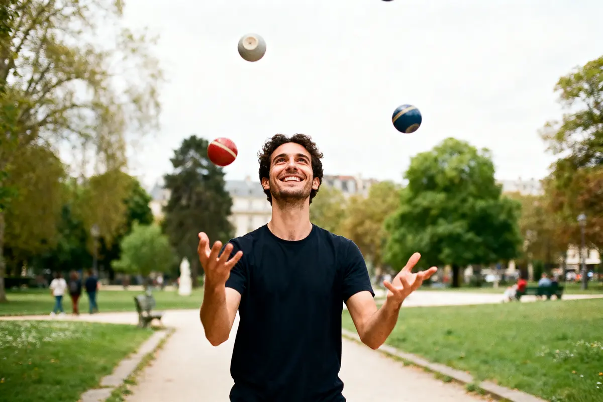 Homme jongle avec quatre balles dans un parc, souriant, entouré d'arbres et de pelouse.
