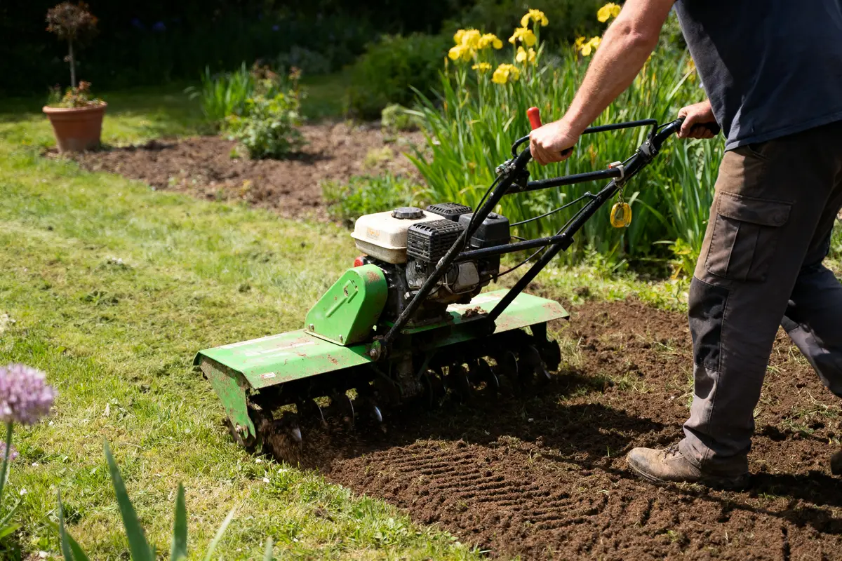 Personne utilisant un motobineur vert pour travailler la terre dans un jardin ensoleillé avec fleurs jaunes.