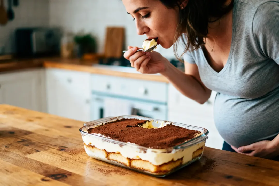 Femme enceinte mange du tiramisu avec une cuillère dans une cuisine, vêtue d'un t-shirt gris.