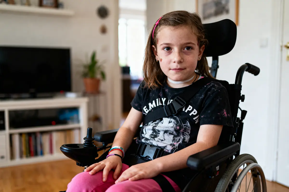 Jeune fille en fauteuil roulant, cheveux châtain, vêtue de rose et noir, dans un salon lumineux.