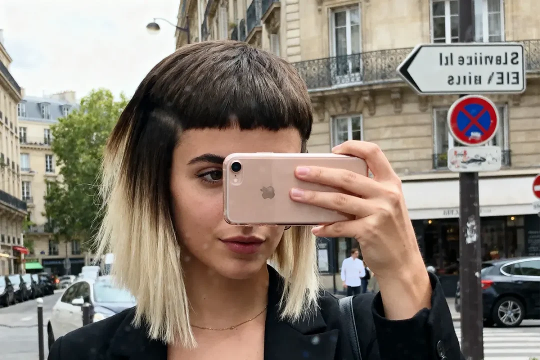 Femme prenant un selfie devant des immeubles parisiens, coiffure originale, smartphone rose, panneau Lavirotte visible.