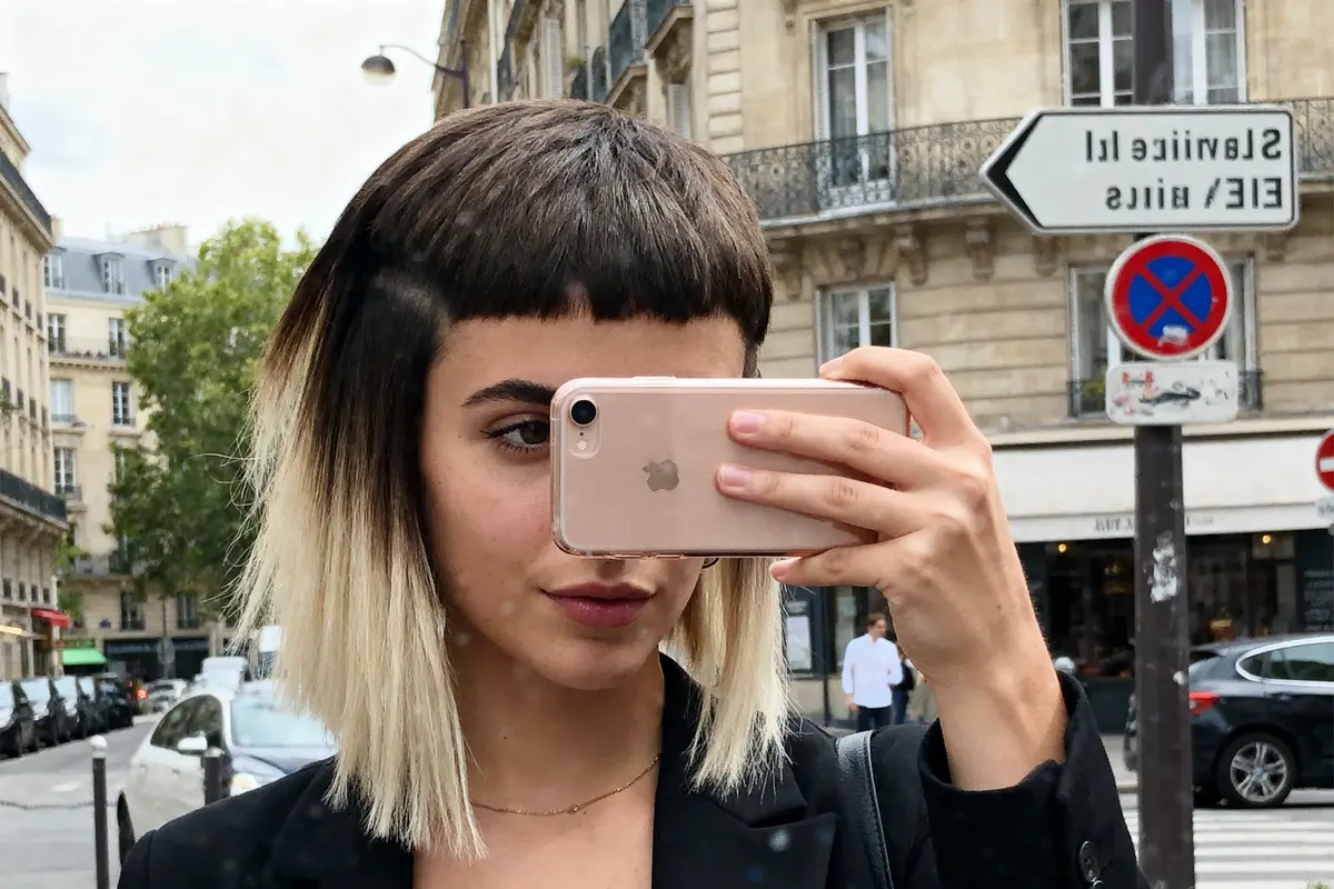 Femme prenant un selfie devant des immeubles parisiens, coiffure originale, smartphone rose, panneau Lavirotte visible.