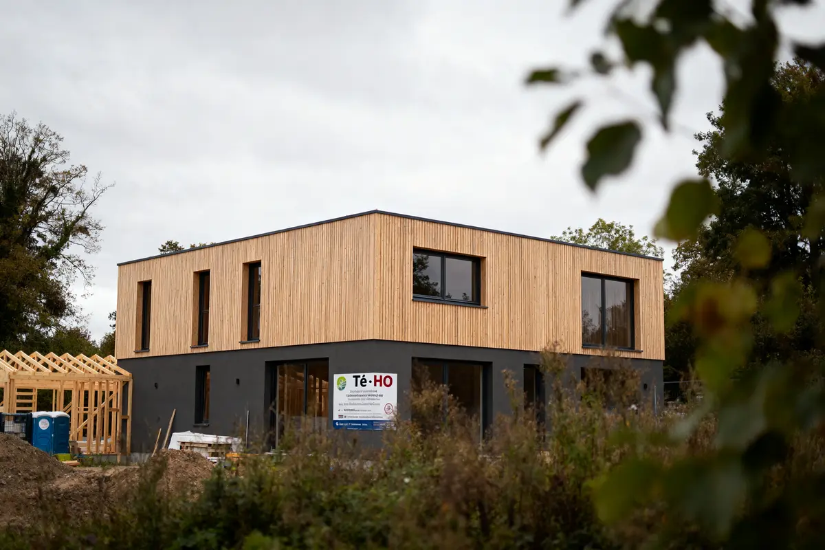 Maison moderne en construction, bardage bois, fenêtres noires, chantier visible, arbres en arrière-plan, ciel couvert.