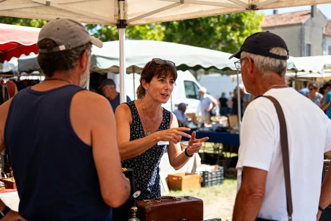 Femme discutant avec deux hommes dans un marché en plein air sous des chapiteaux, objets anciens visibles.