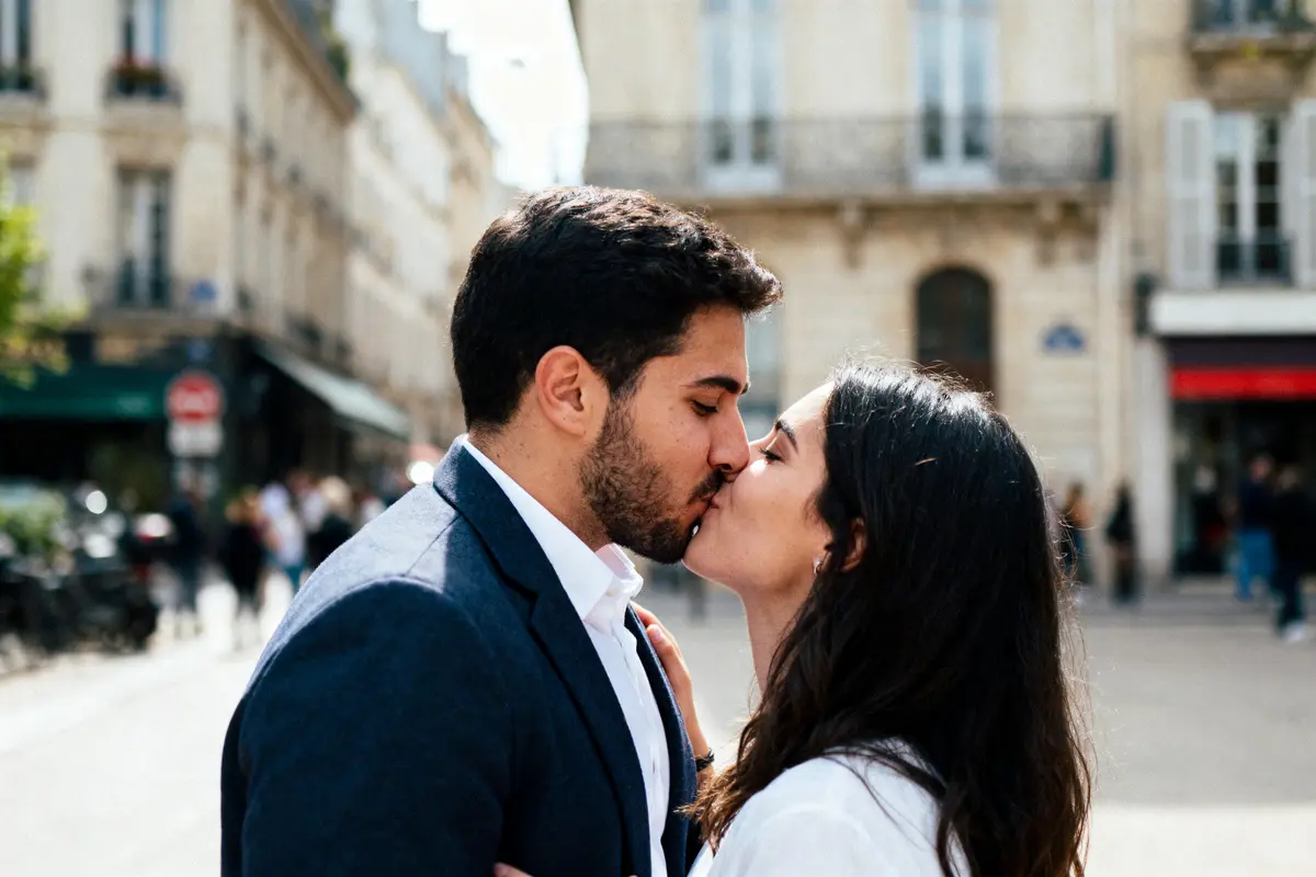 Un couple s'embrasse dans une rue urbaine, sous un ciel clair, entouré de bâtiments historiques.