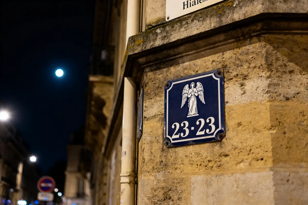 Plaque de rue avec ange, numéro 23-23, ville la nuit, lune visible, bâtiments en arrière-plan.