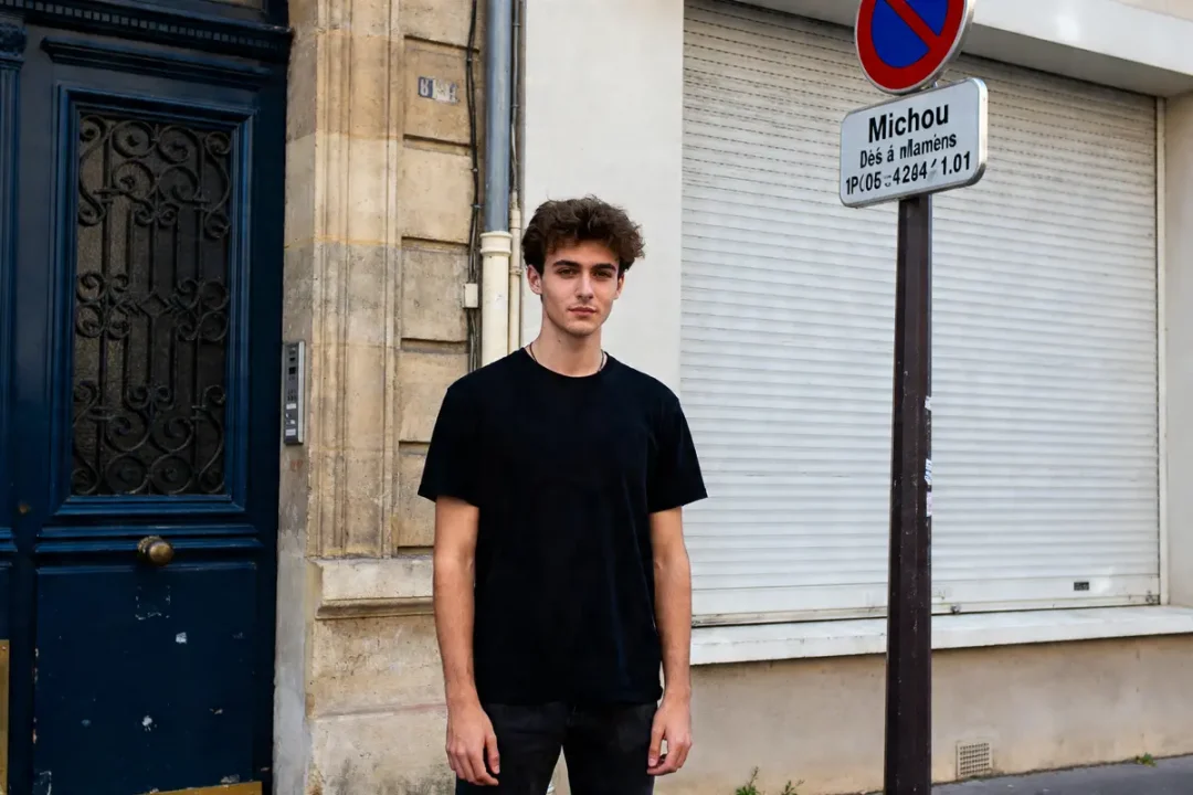 Un jeune homme debout devant une porte et un panneau indiquant l'âge de Michou