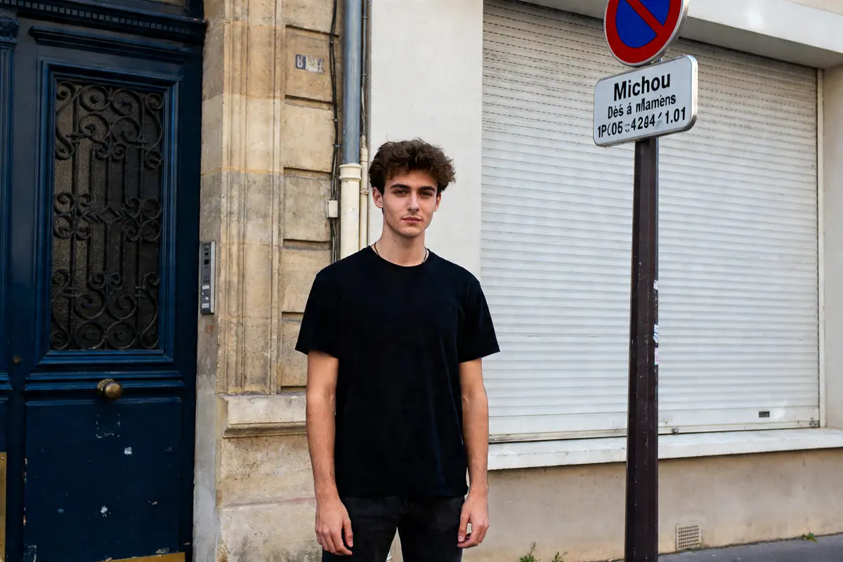 Un jeune homme debout devant une porte et un panneau indiquant l'âge de Michou